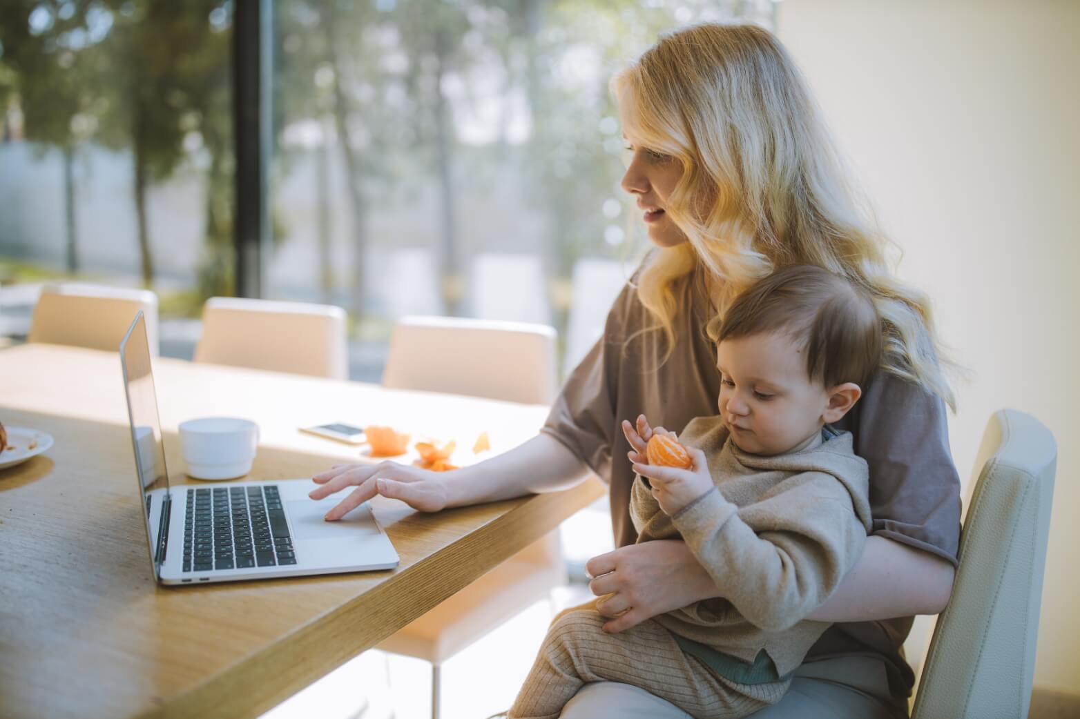Woman working on laptop at table, holding a child with an orange piece.