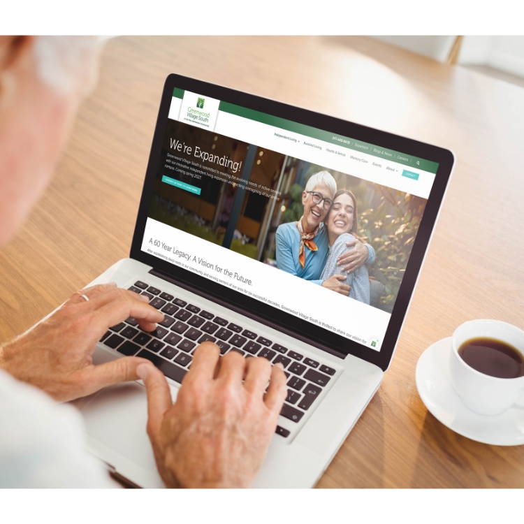 Person browsing senior living community website on laptop at a wooden table with a coffee cup.